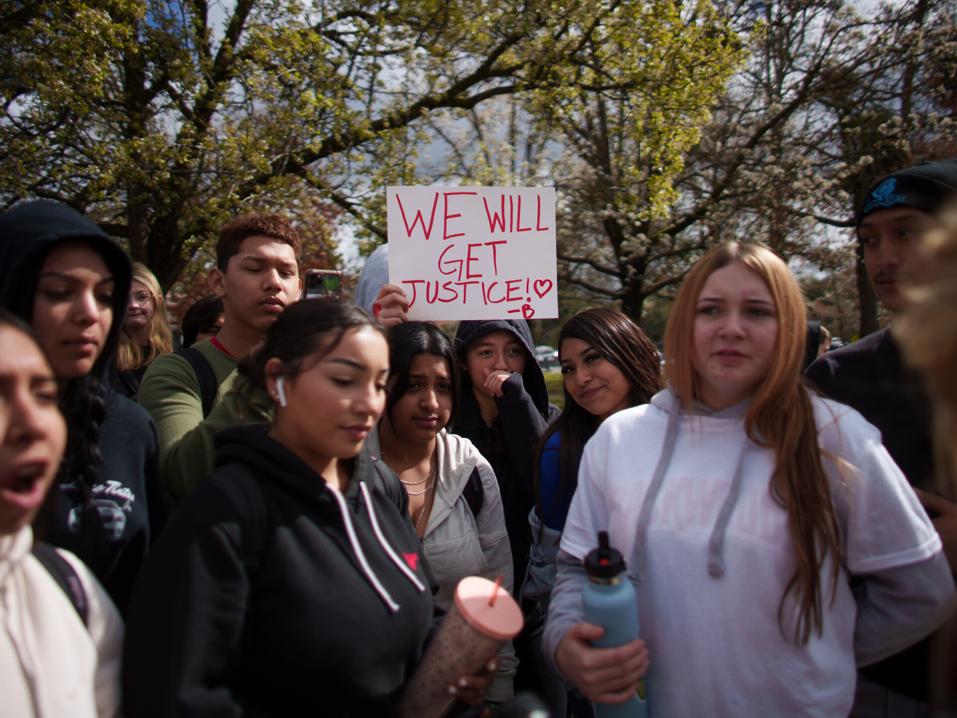 A group of students who appear to be high school aged stand together outside with trees in the background and one visible sign on white paper with red ink, reading 'we will get jutice'