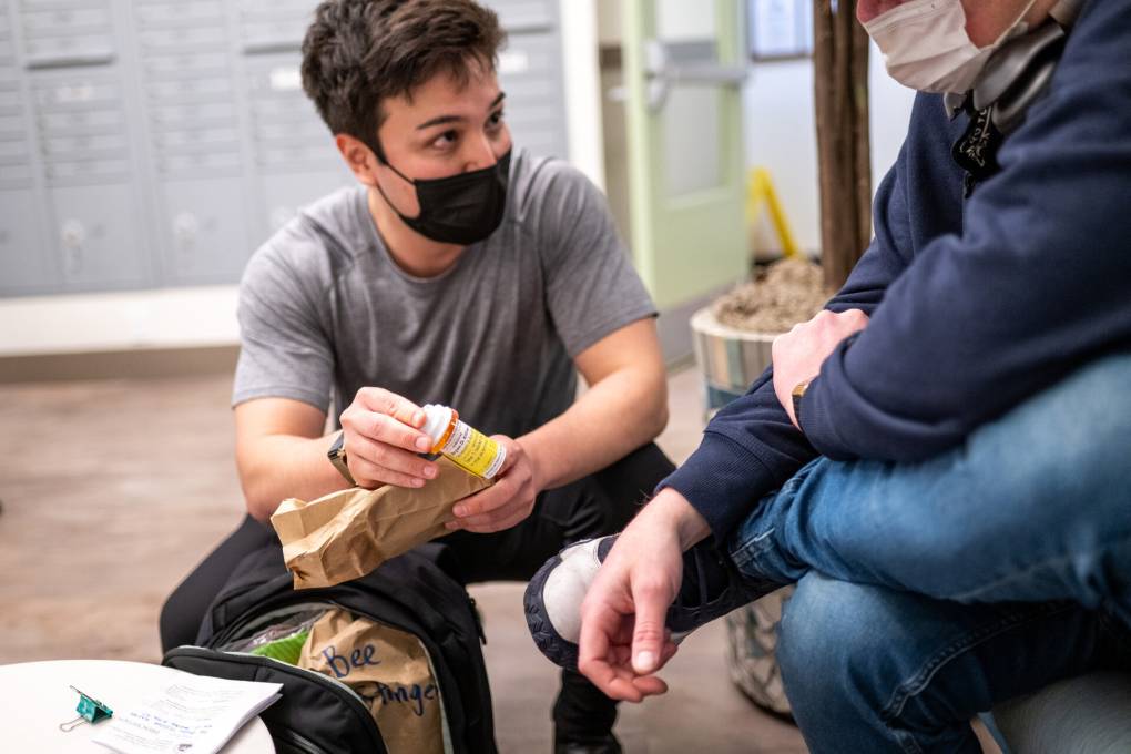 A man wearing a gray T-shirt, black face mask kneels down with an orange medication bottle in hand as he looks up at a patient who is sitting in a chair wearing a navy sweater and jeans.