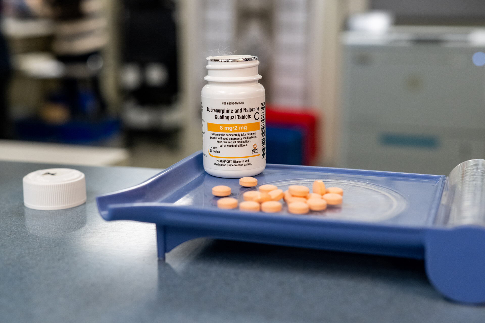 Buprenorphine and Naloxone orange tablets are measured on a blue surface inside a medical clinic. The bottle is white with black and orange letters.