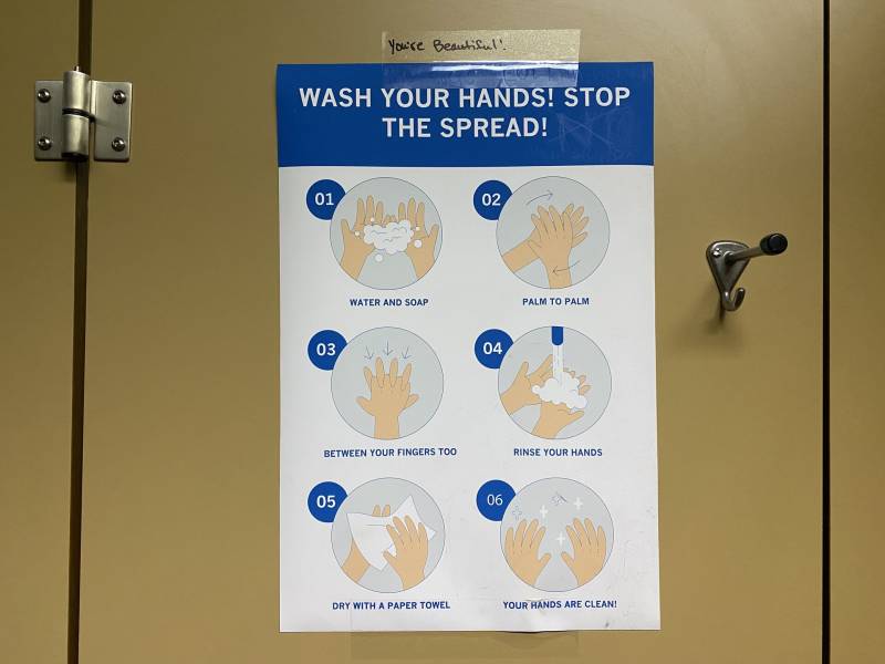 A poster taped to a beige bathroom stall wall that reads WASH YOUR HANDS! STOP THE SPREAD! and gives pictorial instructions for handwashing below