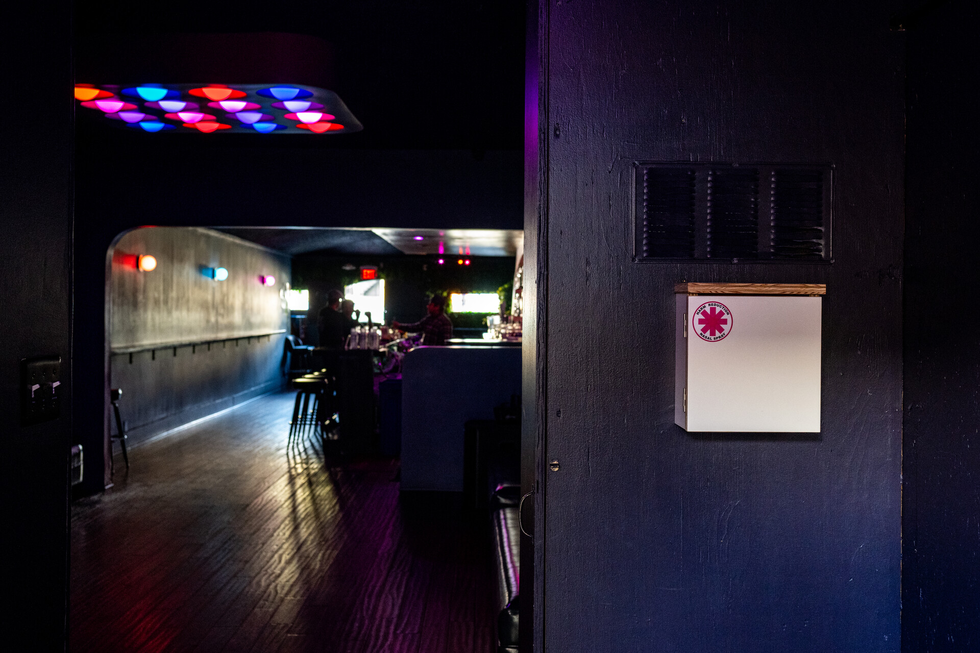 A closed white box with a pink asterisk logo hangs on a wall in a dimly lit bar with a bar counter further back and a bartender and two patrons.