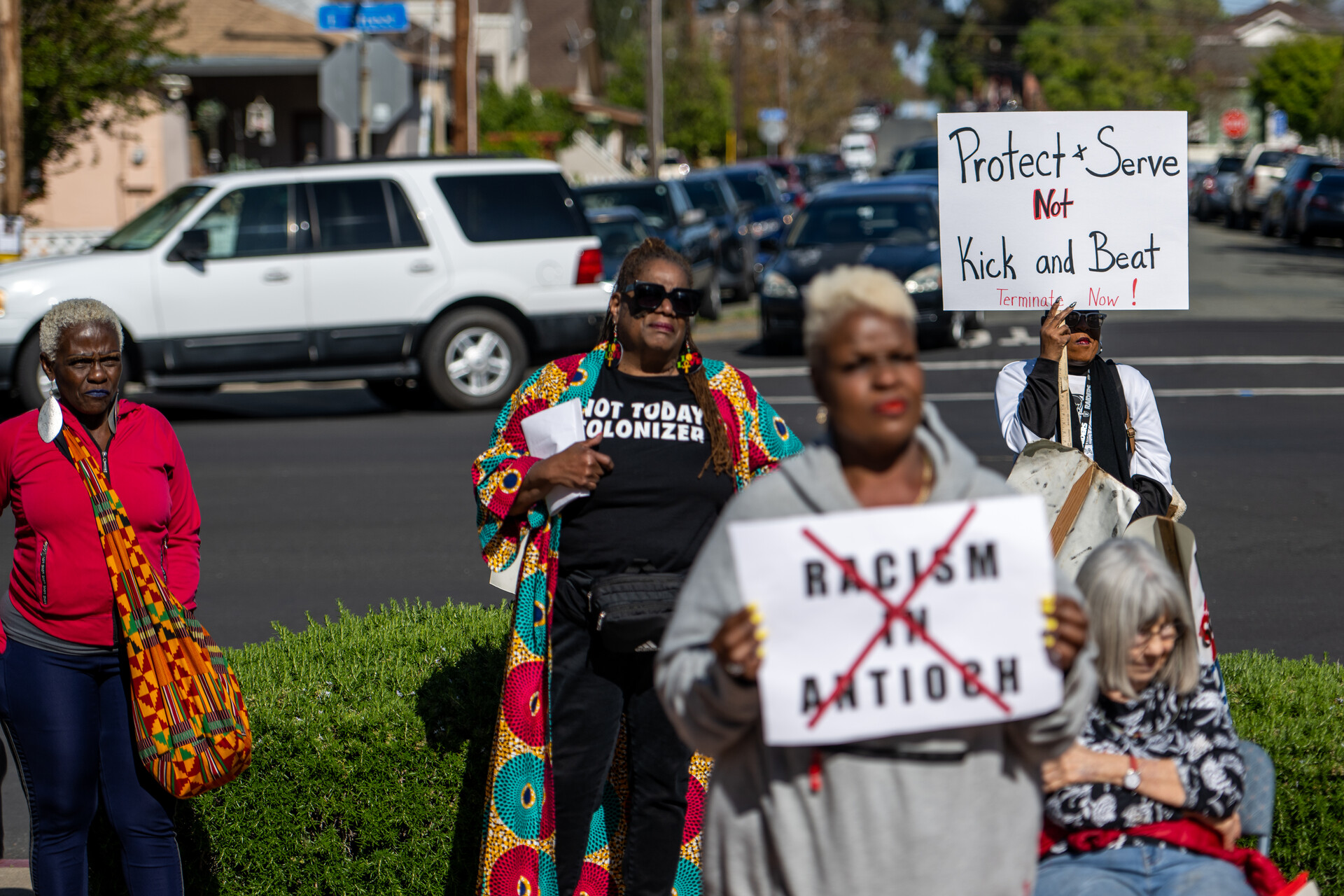 A small group of Black women stand on the sidewalk holding signs and protesting racism among police in Antioch.