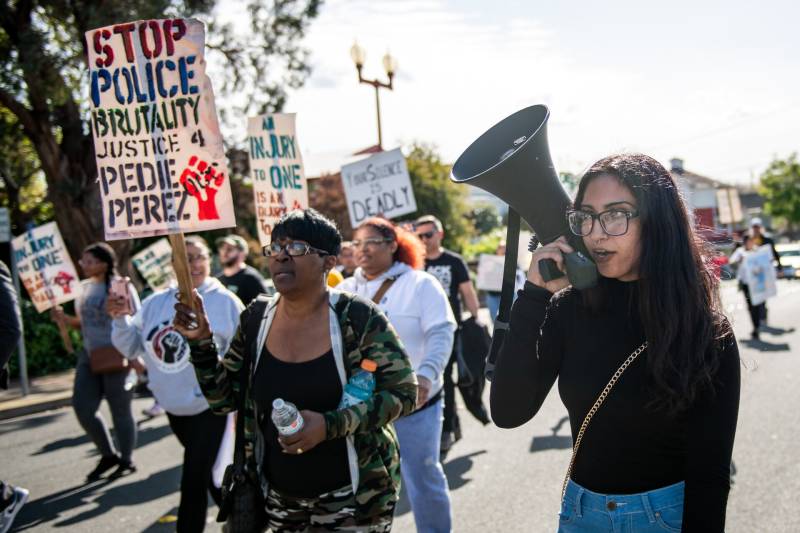 A young woman with a bullhorn leads protesters on the street holding signs.