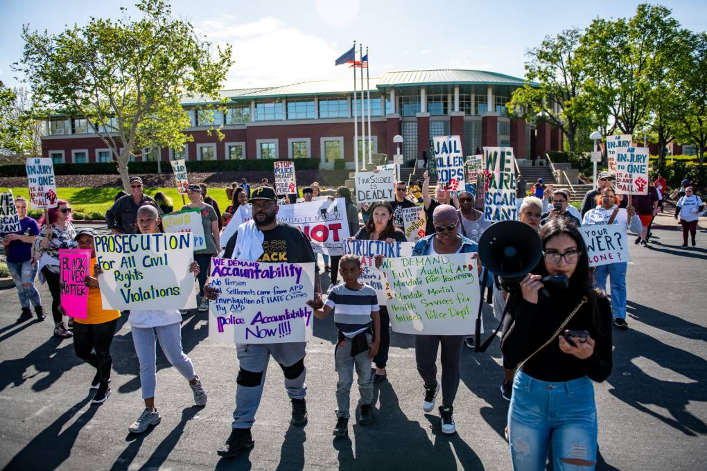 Dozens of people rally in the street in front of an administrative building holding signs signs, following one woman who speaks into a bullhorn.
