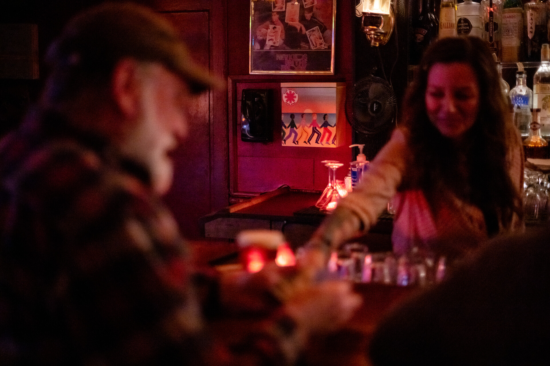 A white woman stands behind a dimly lit bar as an older white man with a white beard speaks smilingly to her