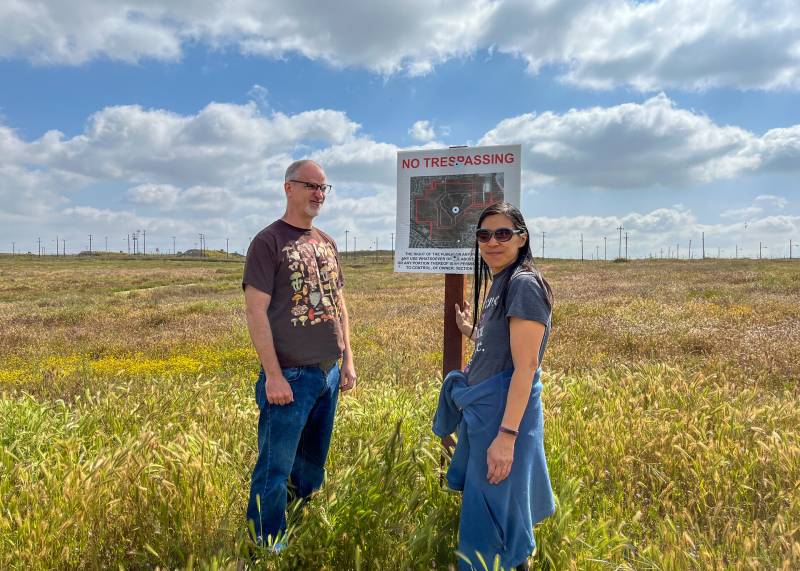 A white middle-aged man and woman, both white, stand in a field with wildflowers, in front of a 'No Trespassing' sign.