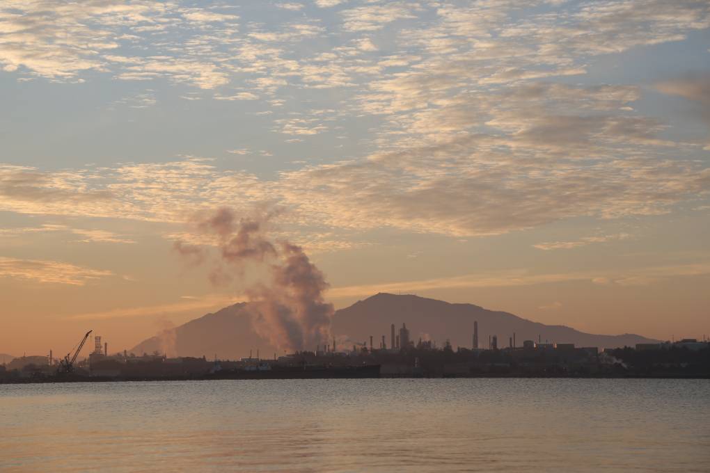 A refinery with smoke rising across a body of water with a mountain in the background around sunset.