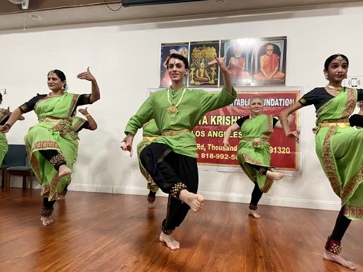 A young mixed raced teenager along with several other people dressed in green attire with brownish stitching on the ends of their clothes perform a dance in a studio.