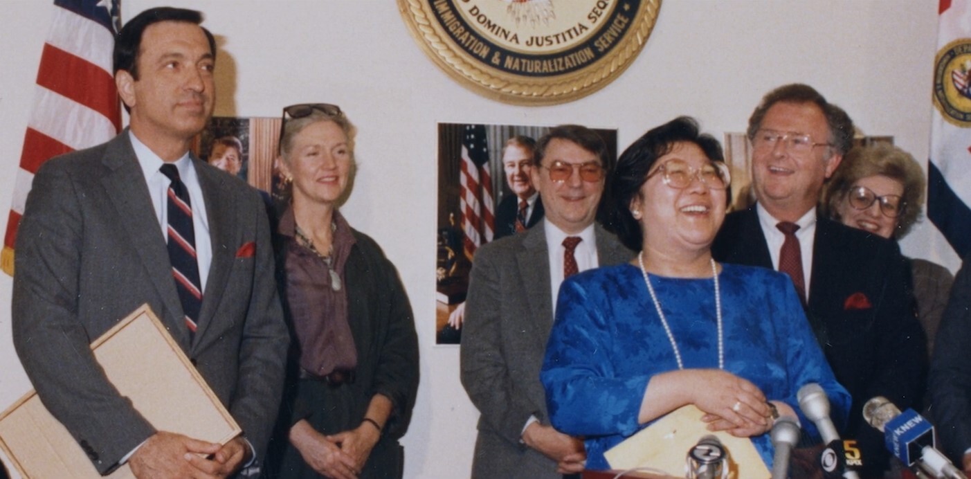 A Chinese woman wearing large, '80s-style glasses, a bright blue dress, and a long strand of pearls, with chin-length black hair, smiles broadly, while others around her, all white, also smile, in a government office in front of a bank of microphones.