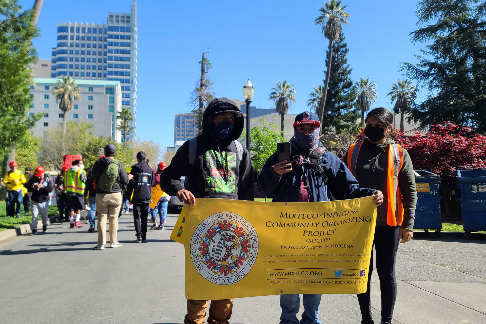 Three demonstrators, all wearing dark sweatshirts and face masks that obscure their faces, hold up a yellow sign with a bright, round insignia that says Mixteco/Indigeni Community Organizing Project, standing in the middle of a street with trees and high-rise buildings beyond them, on a blue-sky day.