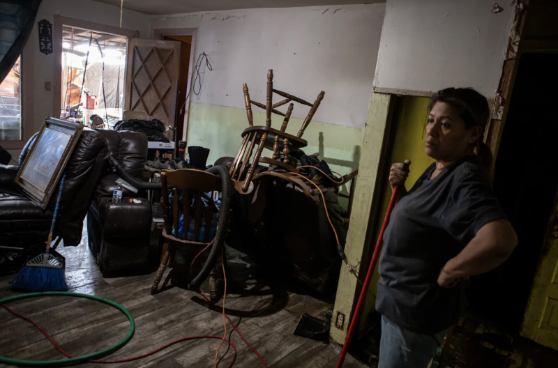A woman holds a broom while looking at a room with chairs and other materials stacked up.