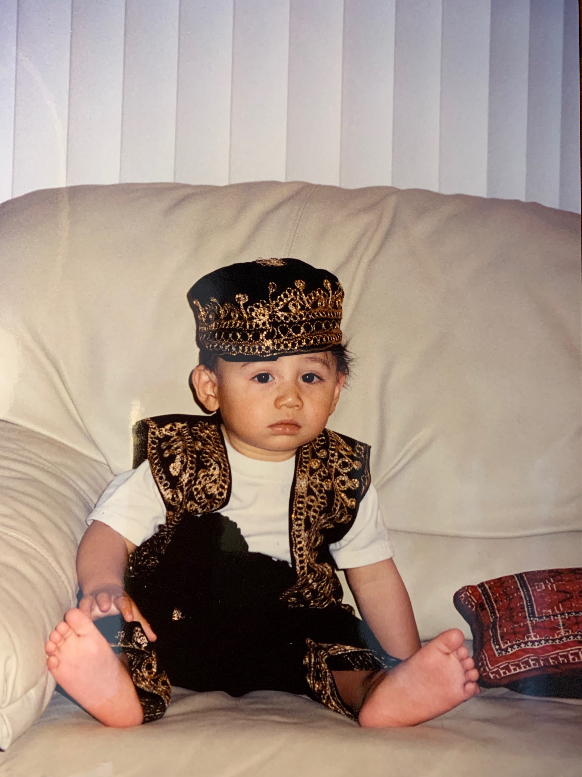 A cute baby is seen sitting barefoot on a white, leather sofa. He wears black and gold, traditional Lebanese garb.