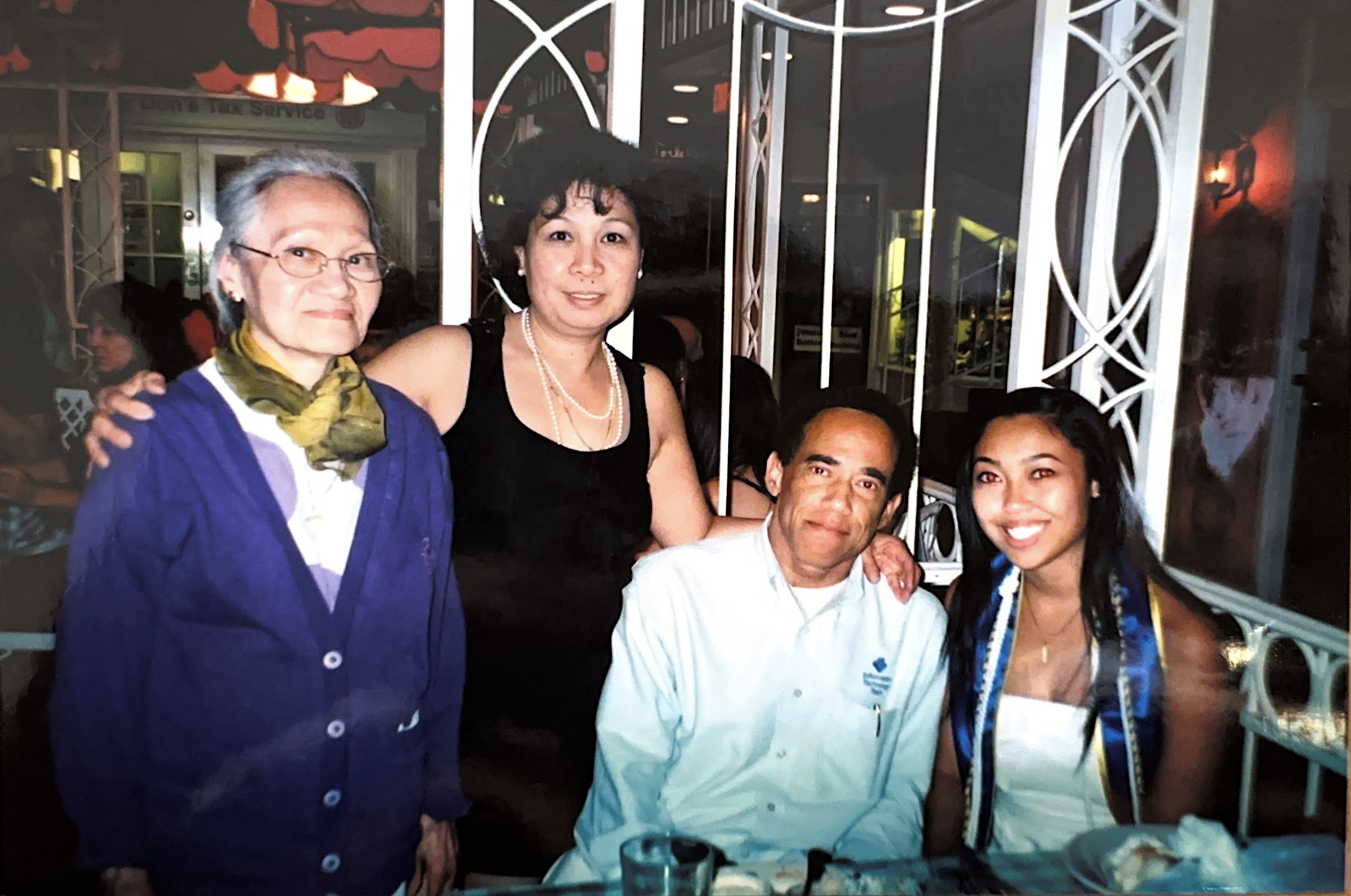 A family is pictured sitting inside a restaurant setting. A grandmother, two parental figures, and their young daughter all smile for the camera. The daughter wears a royal blue college graduation sash around her shoulders.
