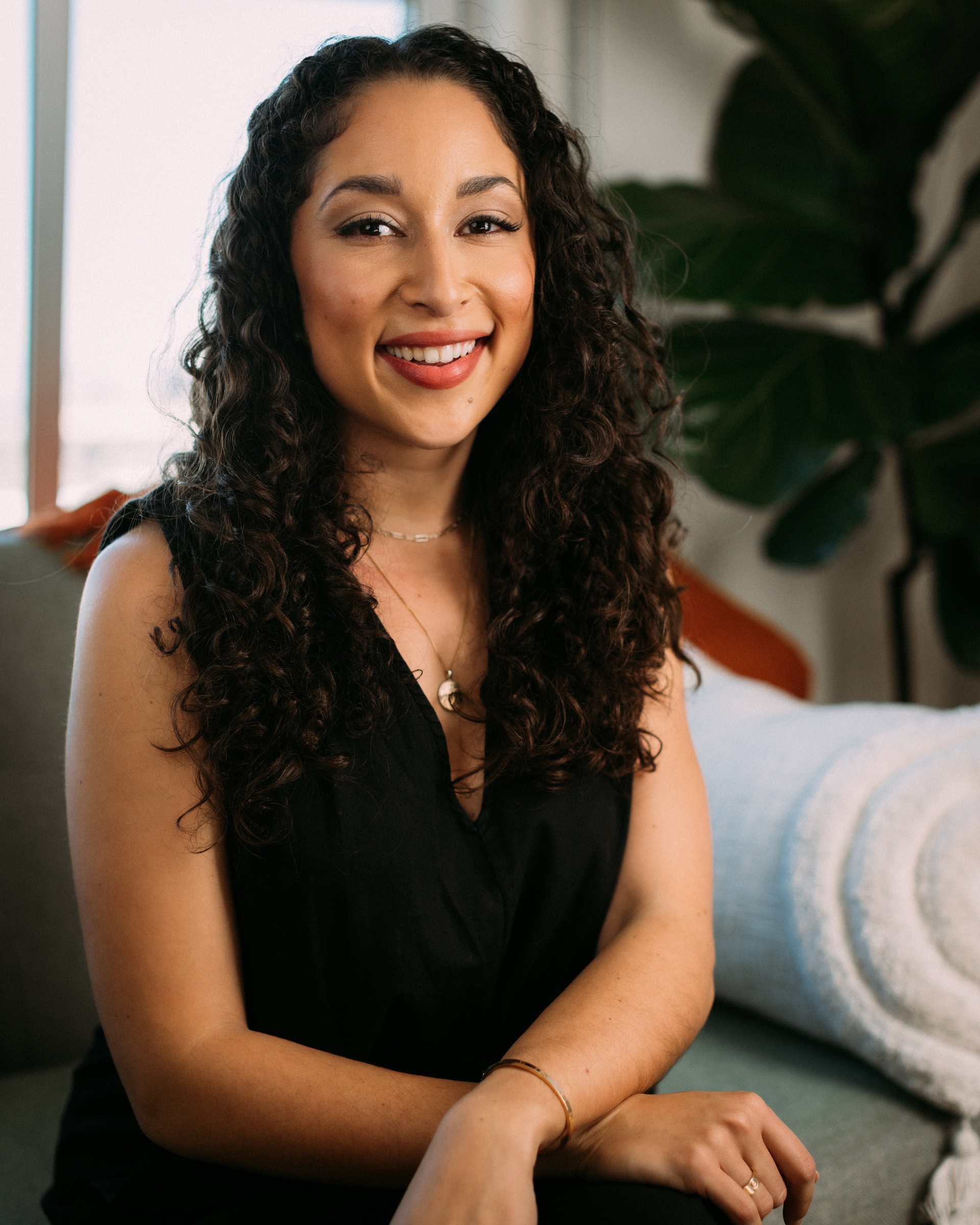 A woman smiles from a gray sofa. She has long, curly brown hair and a friendly face. She wears a gold necklace and a black, sleeveless dress. A happy, green house plant is positioned behind her and the light shines brightly on her face.