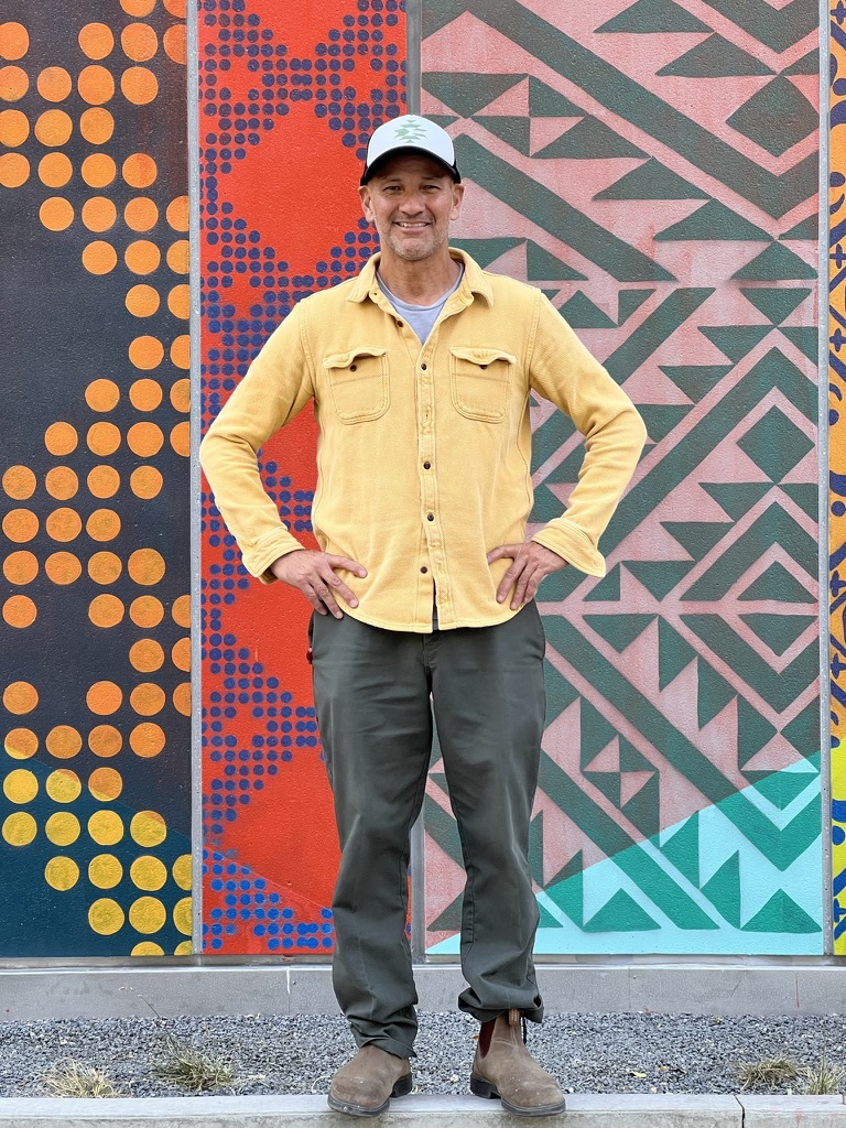 A man with a bright, yellow, long-sleeved shirt smiles in front of a multicolored, funky-patterned mural. He stands with his hands on his hips.