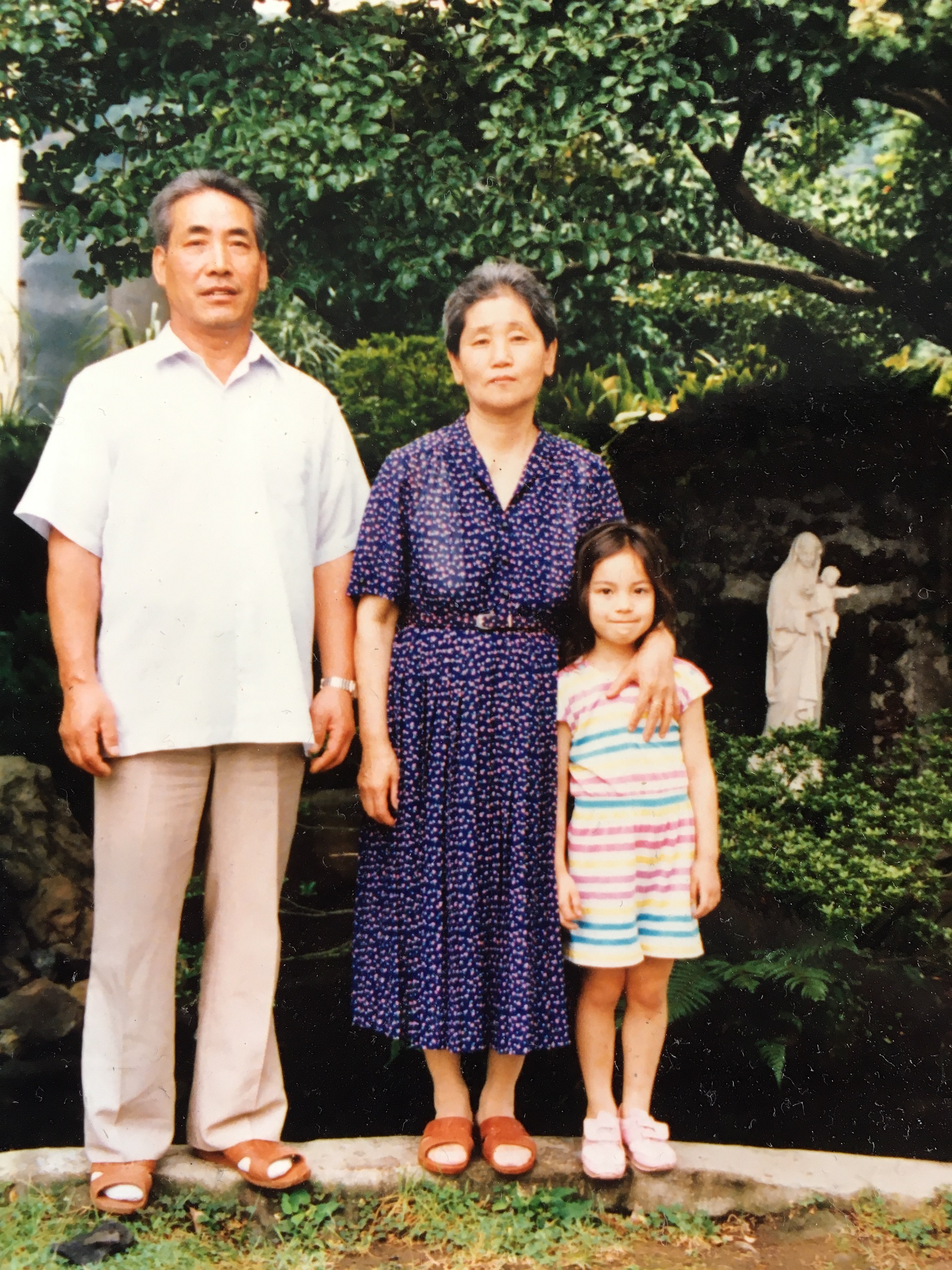 Two grandparents stand with their young granddaughter amid green trees and a pond of water. A ceramic statue of a saint is also in the background. The photo is old and has a classic patina to it.
