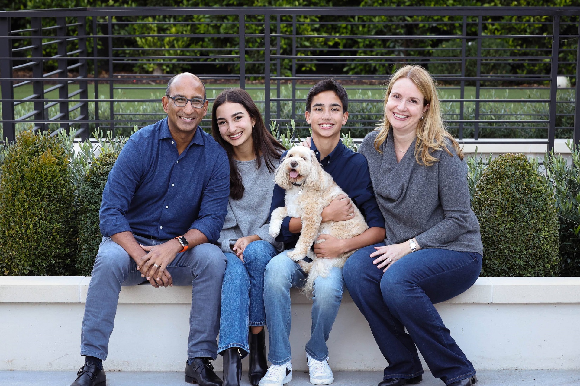 A smiling family of five sit on an outdoor planter with chubby, green bushes behind them. From left to right: A bald dad with glasses sits next to his daughter with long, brown hair and jeans. She, sits next to her brother who smiles holding a happy tan dog with floppy ears. He is seated next to his mother with blond hair and a gray, scoop-neck blouse.