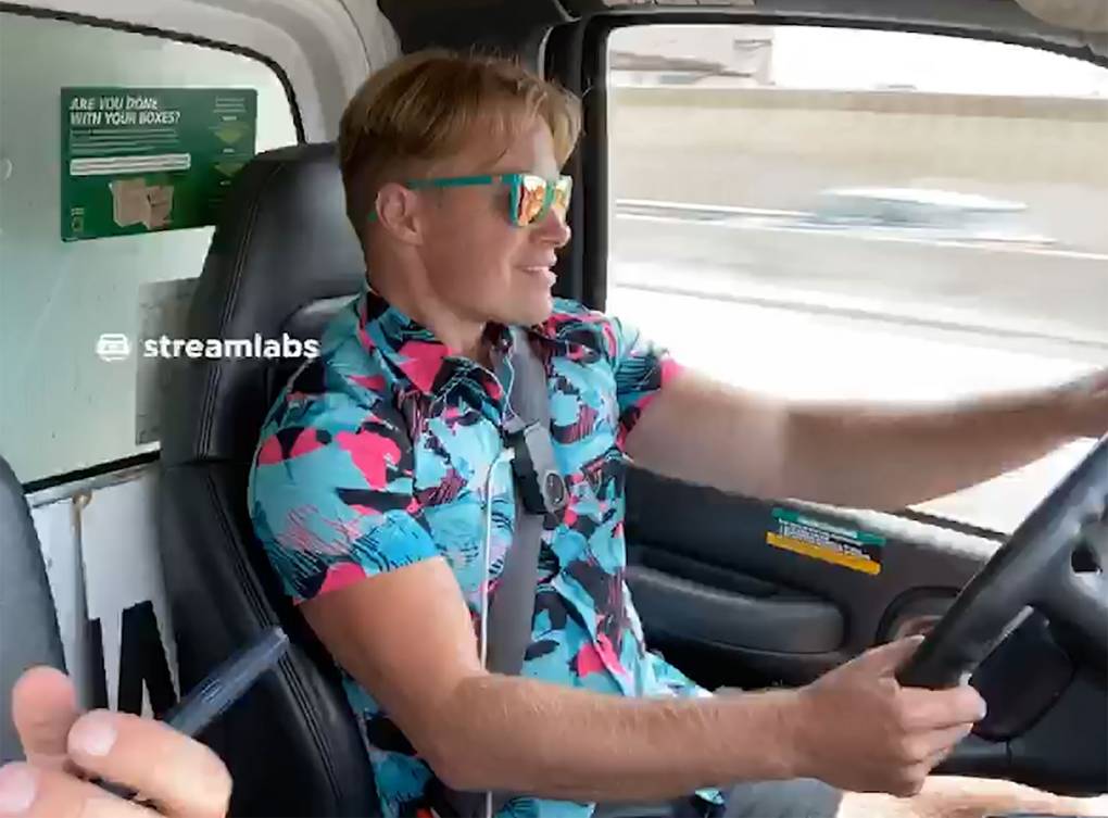 A blonde white man in a bright blue and pink shirt sits behind the wheel of what appears to be a van while driving