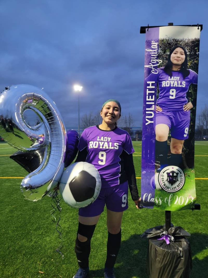 A teenage girl wearing a soccer uniform stands on the field next to a poster of herself. She is holding balloons, including a large number 9, the number on her jersey.