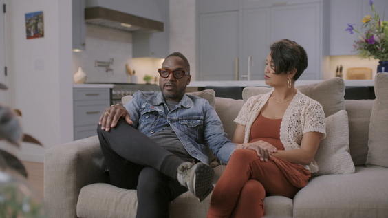A still image of a Black man and an Asian woman sitting on a couch.