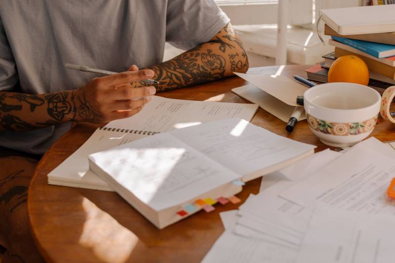 A person with medium-toned skin sits at a wooden table writing on a notepad, surrounded by books. They are wearing a gray t-shirt and have tattooed forearms. We can't see their face.