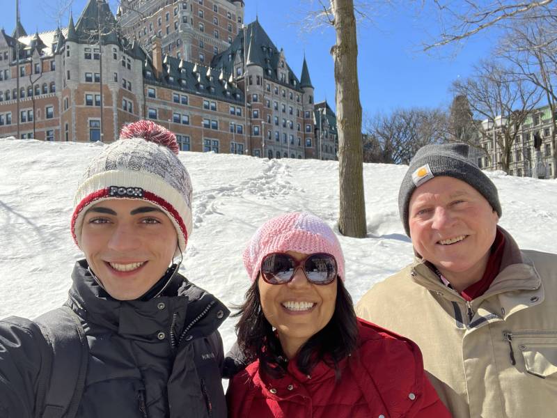 A photo of a mixed race teenager with his parents, a white man and South Asian woman wearing winter clothing outdoors with snow and buildings in the background.