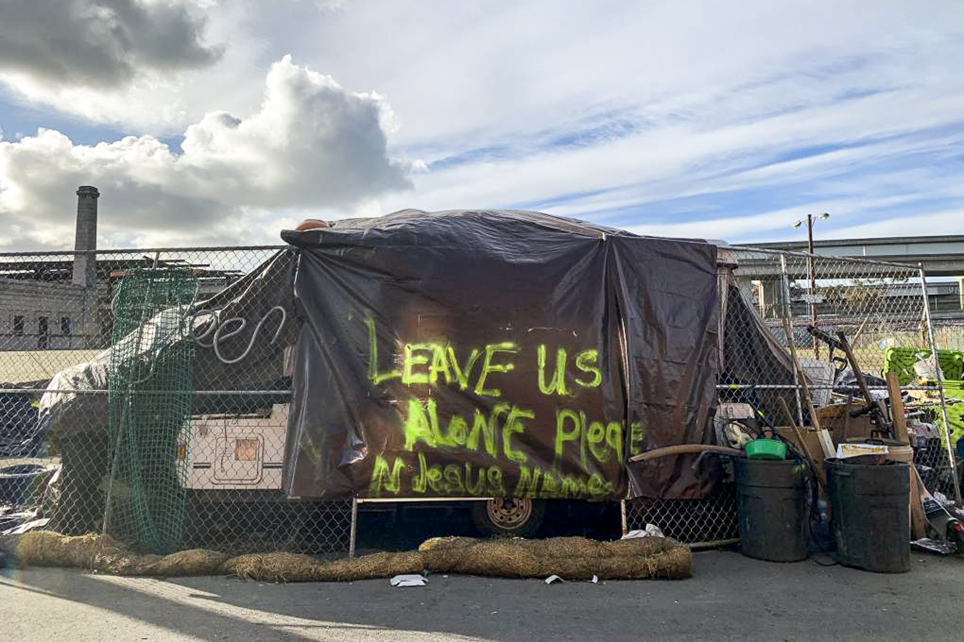 A brown tarp hangs over a trailer with spraypainted letters in yellow reading, "Leave us alone in the name of Jesus."