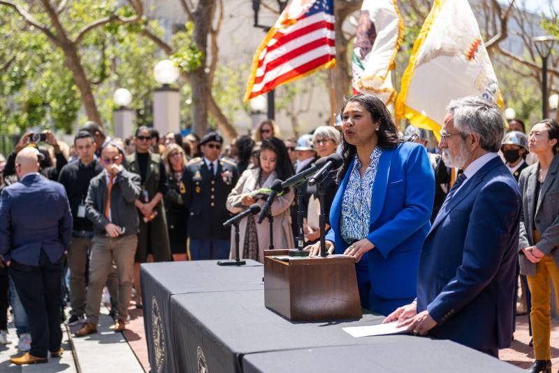 Mayor London Breed speaks outside at a lectern, next to a bearded white man, as a large crowd looks on.
