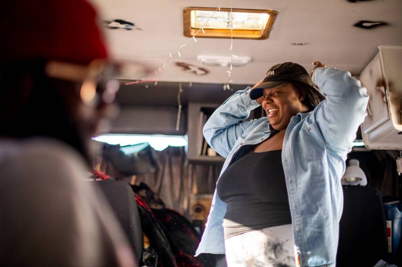 An African American woman with a black shirt and a denim jacket fixes the baseball hat on her head inside her RV as she laughs.