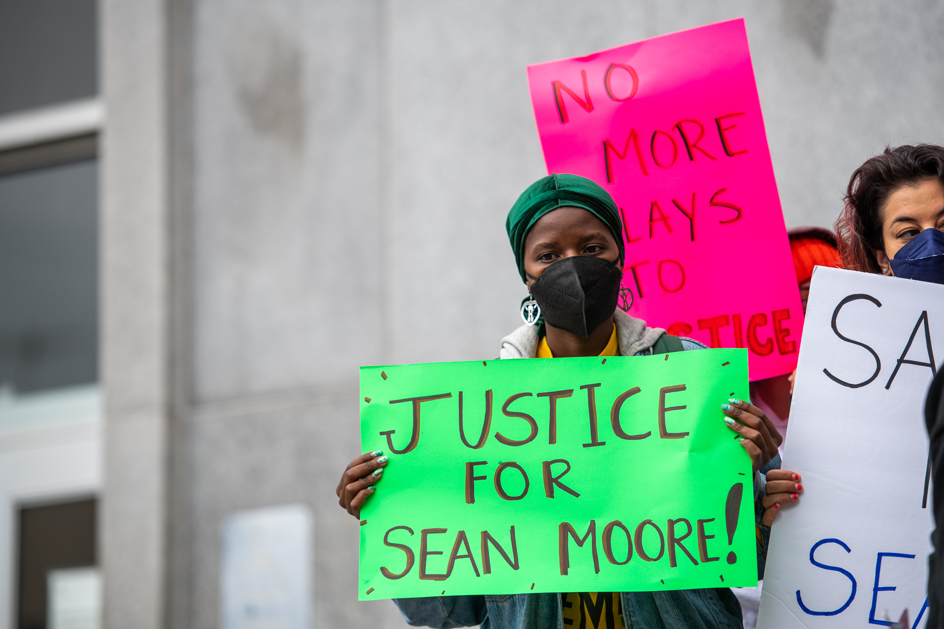 A woman with a green headscarf and black face mask holds a green sign that reads, "Justice for Sean Moore!"