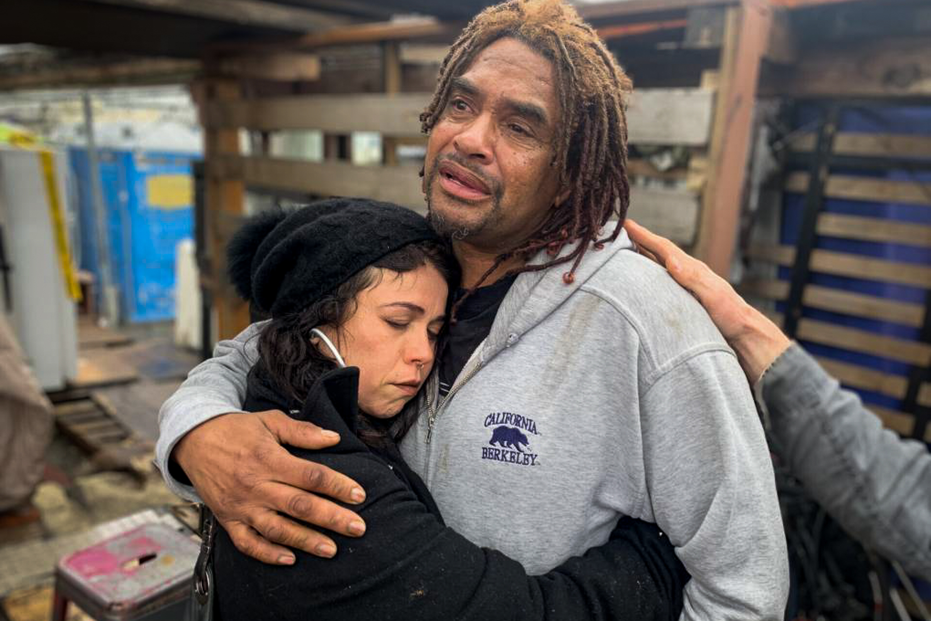 A woman in a black beanie and jacket hugs a man who is crying wearing a gray, hooded sweatshirt.