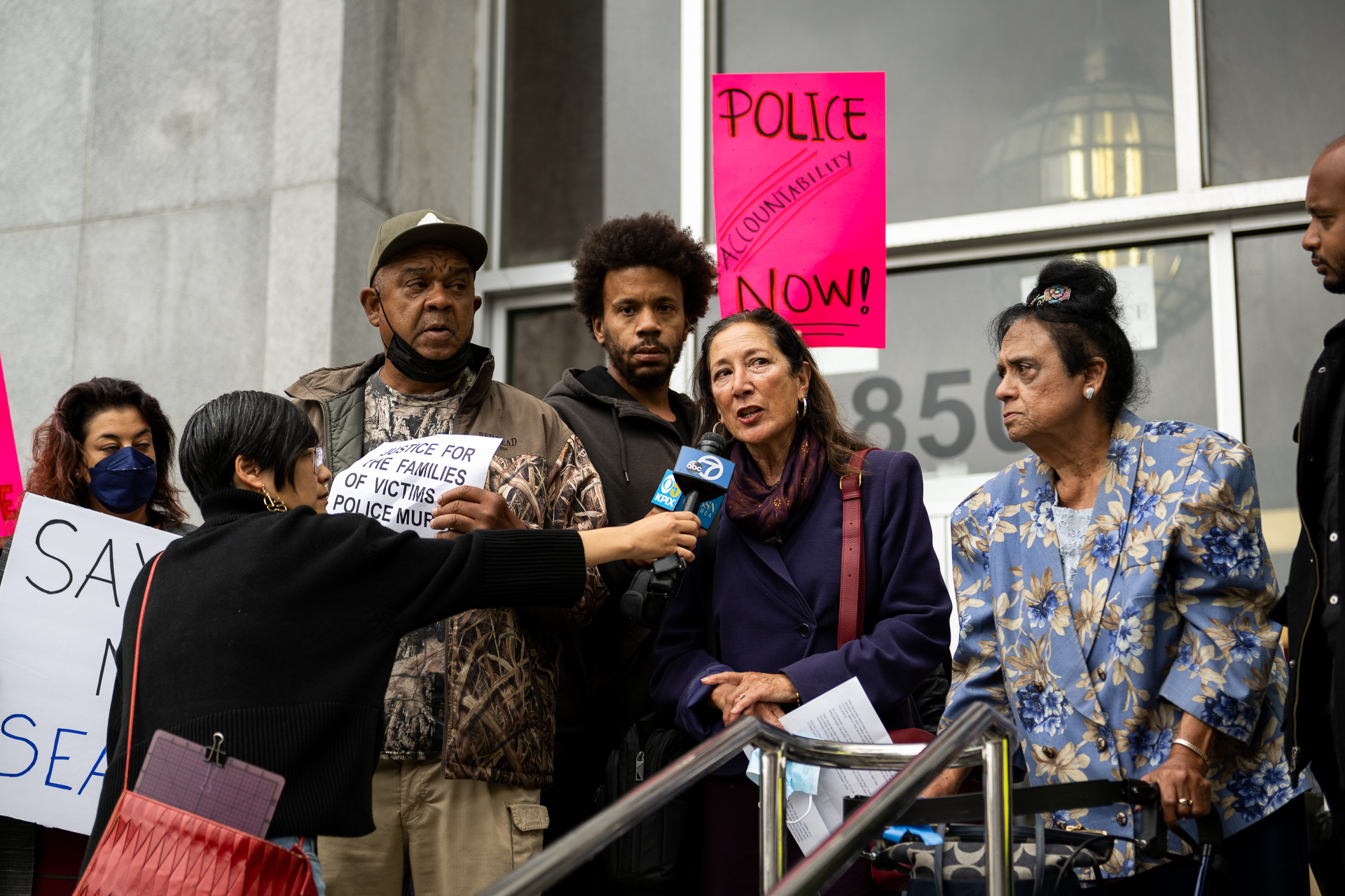People gather on the steps of the Hall of Justice in San Francisco. A news reporter holds out an ABC7 microphone toward a woman with shoulder-length, brown hair and a navy coat on. Many people surround her. Some people hold signs that read, "Police accountability now!"