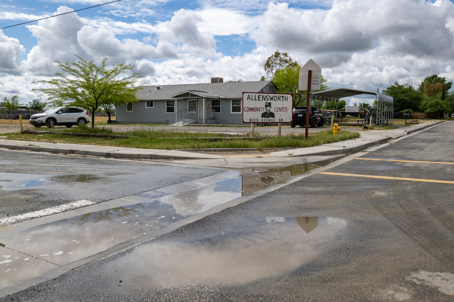 A gray building with a sign out front that reads, "Allensworth Community Center." A white SUV is parked in the driveway and gray clouds hover above. The road surrounding the property is visibly wet from flooding.