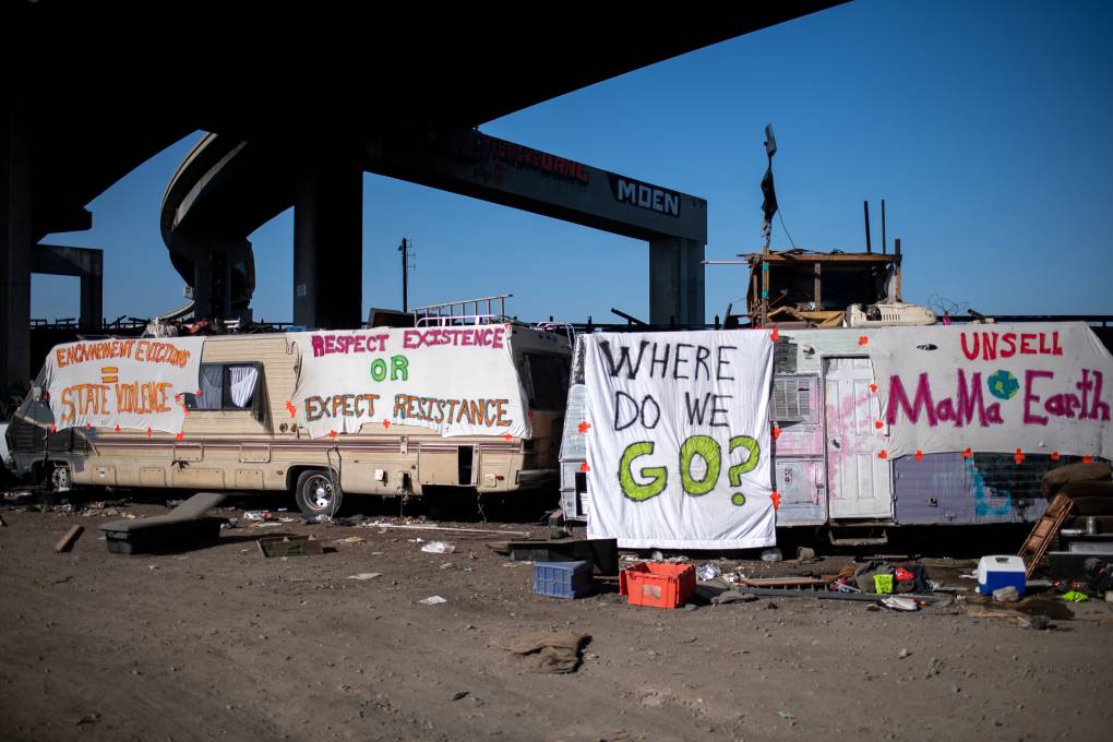 RVs in an encampment under a freeway ramp with signs that say 'Where do we go?' draped on the nearest RV.