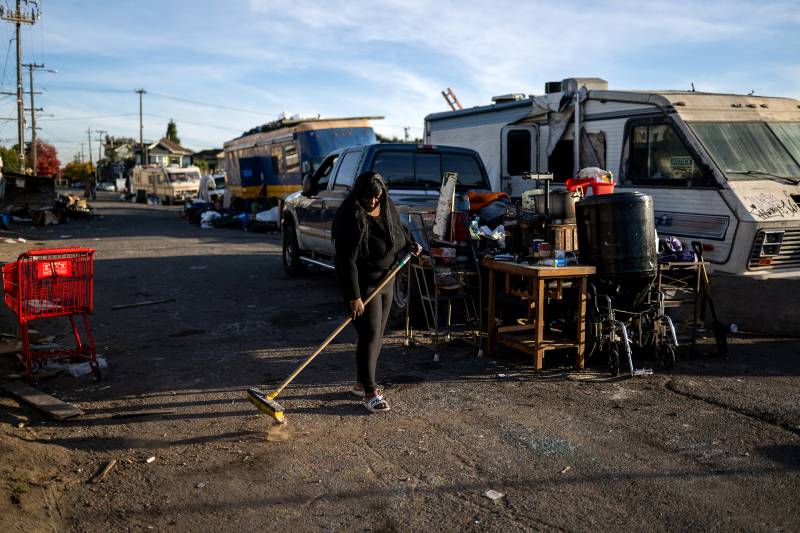 An African American woman sweeps a dirt path with a broom with cars and RVs parked behind her.
