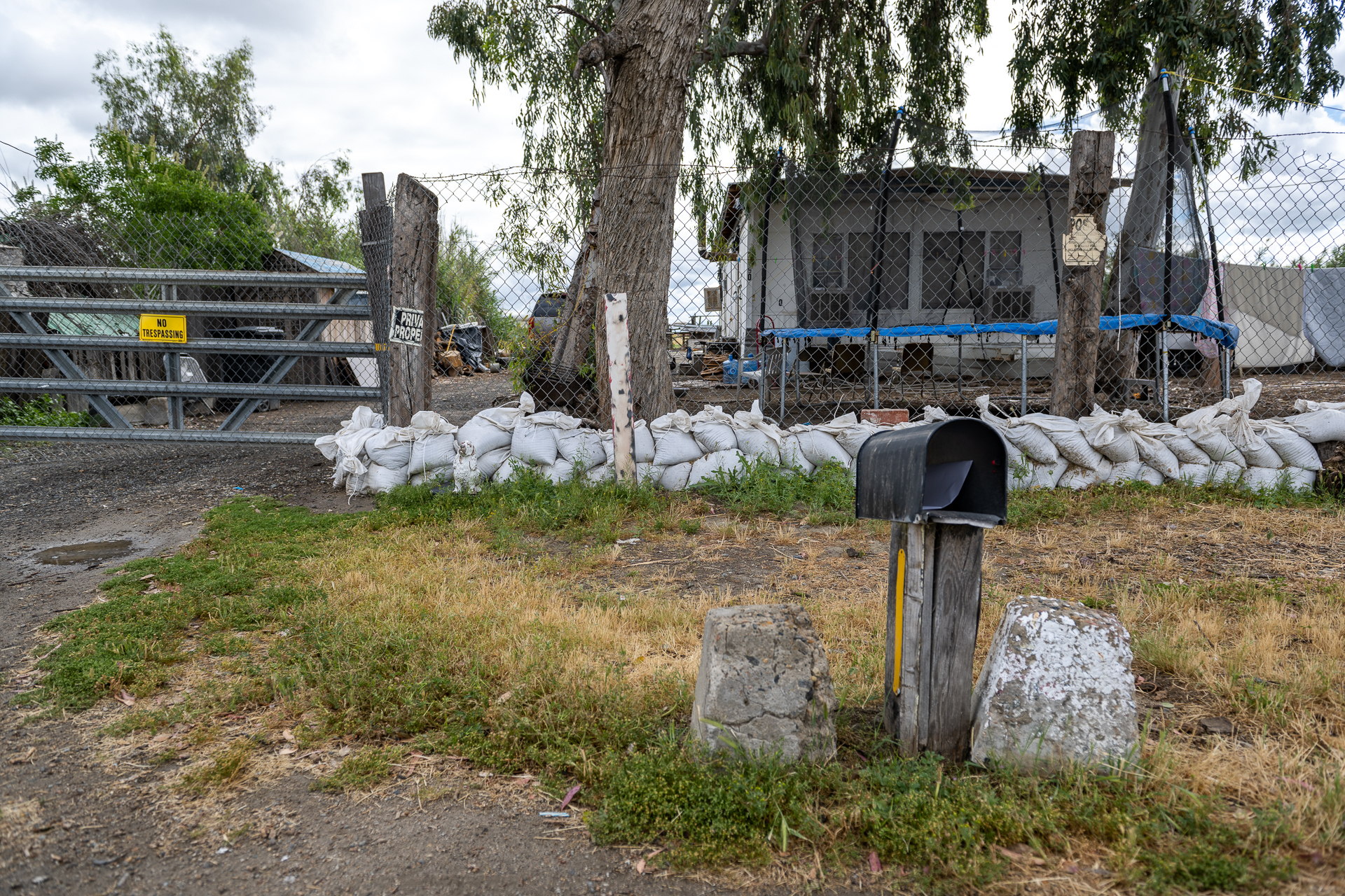 A pile of sandbags line the perimeter of a small home. In the front yard, a blue trampoline is visible and a weathered, black mailbox sits on top of a thick piece of wood.
