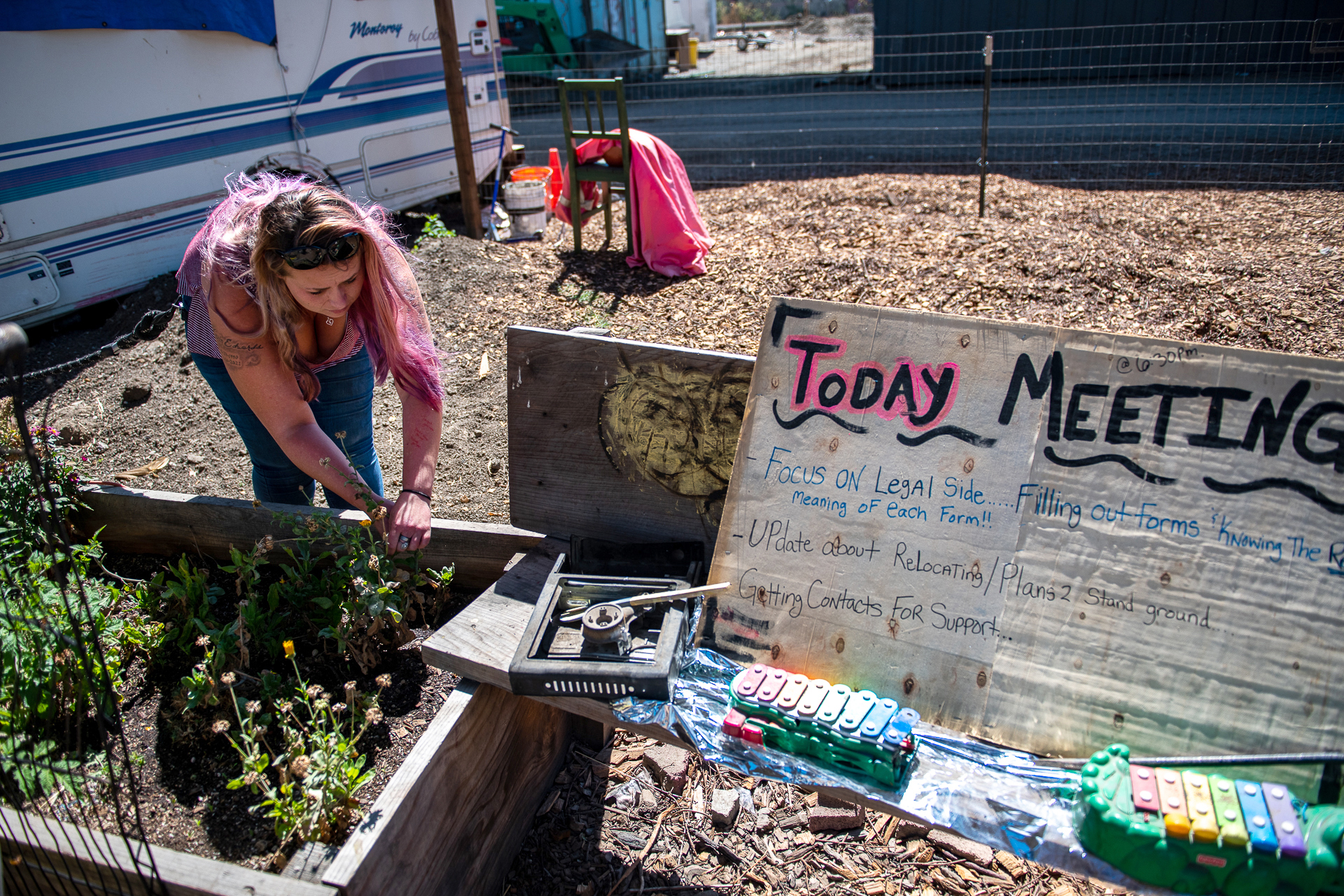 A woman in a pink T-shirt with blonde hair pulls weeds from a planter box that holds a sprouting garden. Little orange flowers are blossoming. A nearby white board reads, "Today Meeting." A trailer is seen to the left in the background.