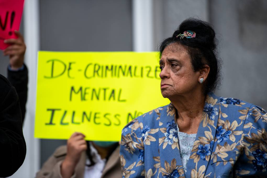 An older woman with a somber face wears a blue blazer with pastel flowers on it. She looks to the left. A yellow sign behind her reads, "Decriminalize mental illness now!"