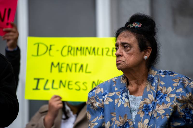 An older woman with a somber face wears a blue blazer with pastel flowers on it. She looks to the left. A yellow sign behind her reads, "Decriminalize mental illness now!"