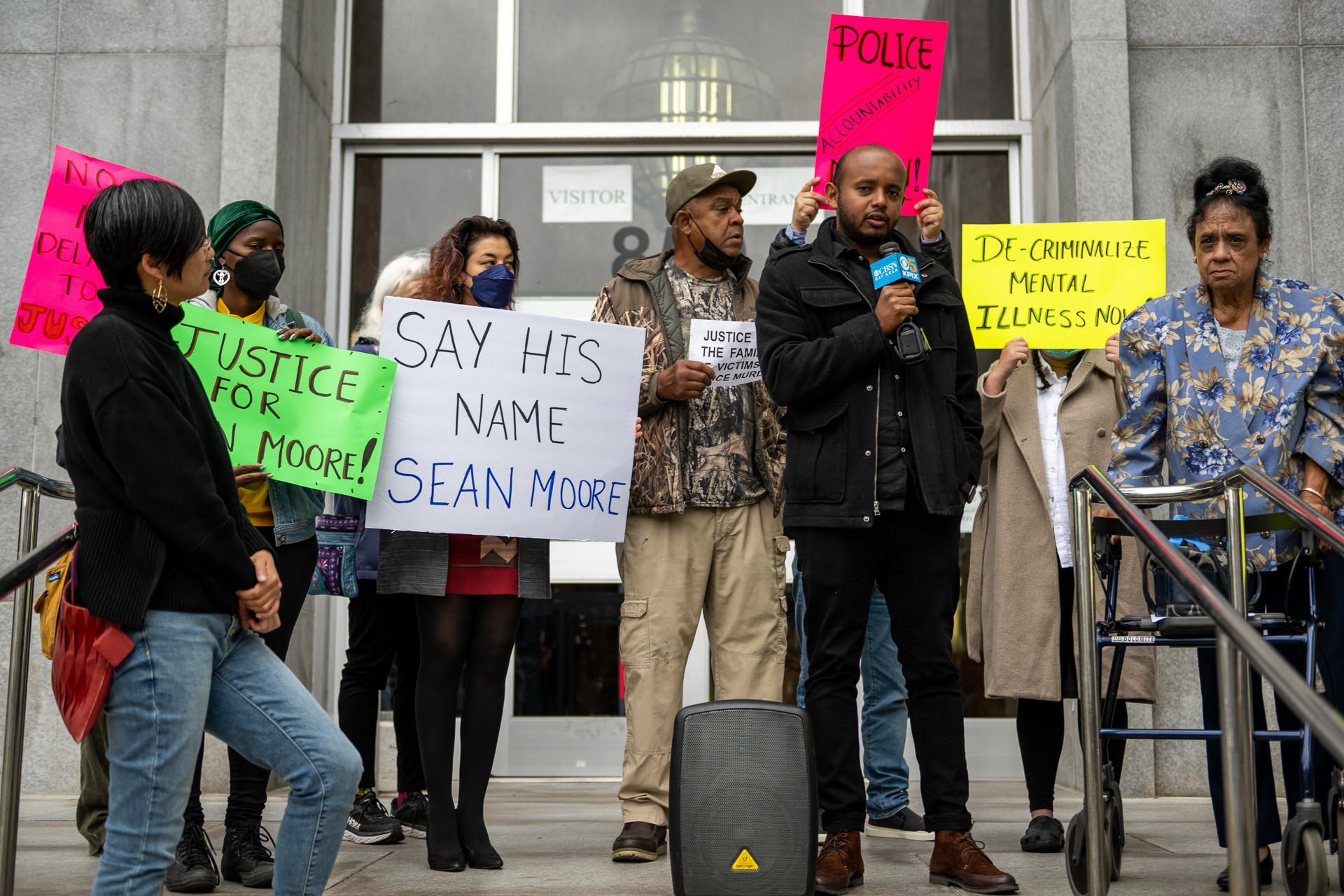 A group of people are gathered on the steps of the Hall of Justice in San Francisco. One woman is behind a walker. Many have signs in their hands that read, "Justice for Sean Moore," "Say his name Sean Moore."