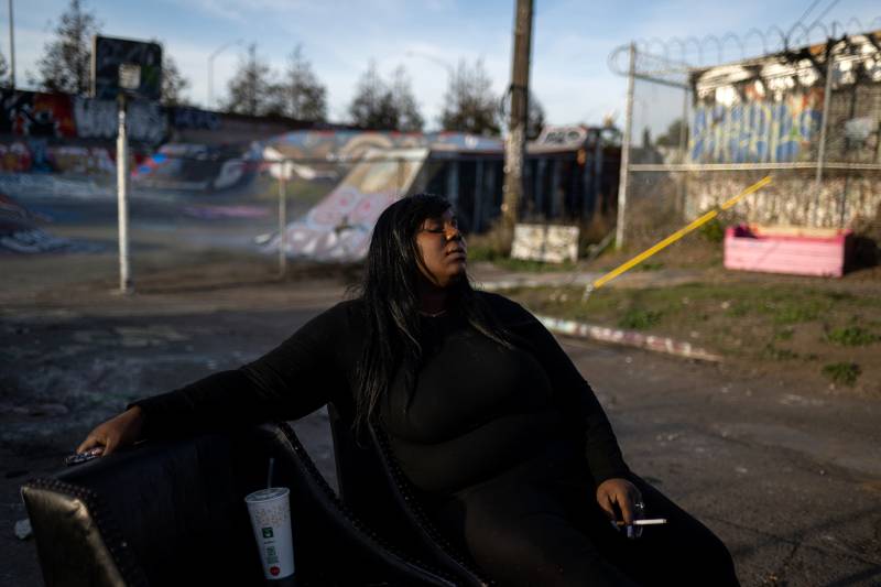 An African American woman sits on a chair outside by the road, closing her eyes as she smokes a cigarette, wearing a black outfit.