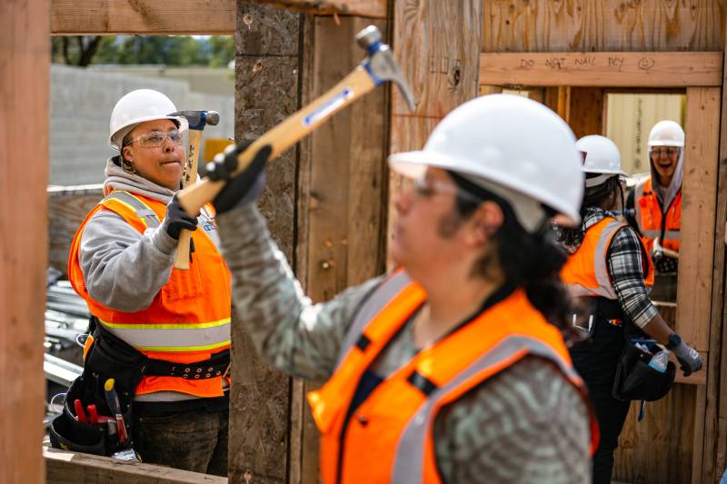Two women wearing construction uniforms hold hammers near wooden boards at a construction site.