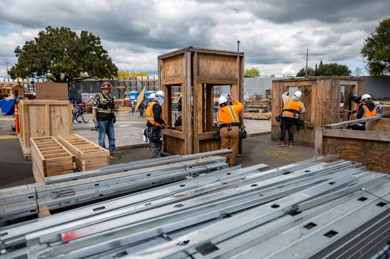 A group of people wearing construction equipment hold a wooden structure as one person looks on while another works on a different wooden structure.