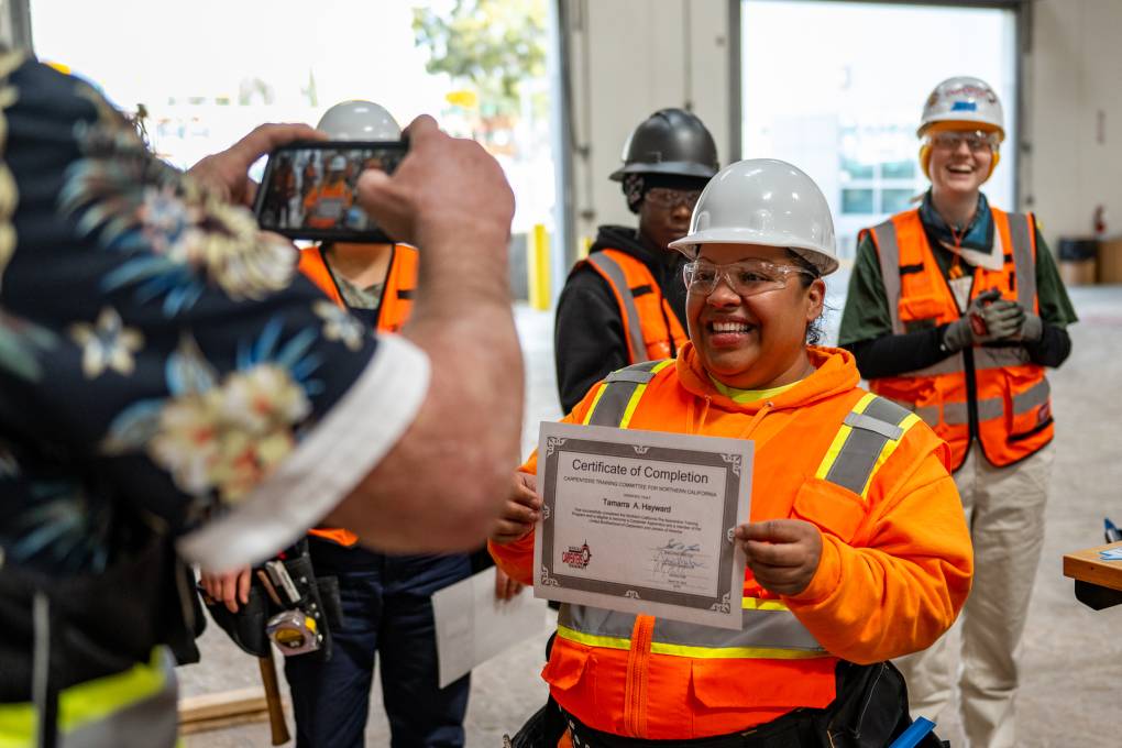 A woman wearing a construction uniform poses with a certificate in both hands as a person takes a picture with other people in construction uniforms stand behind her.