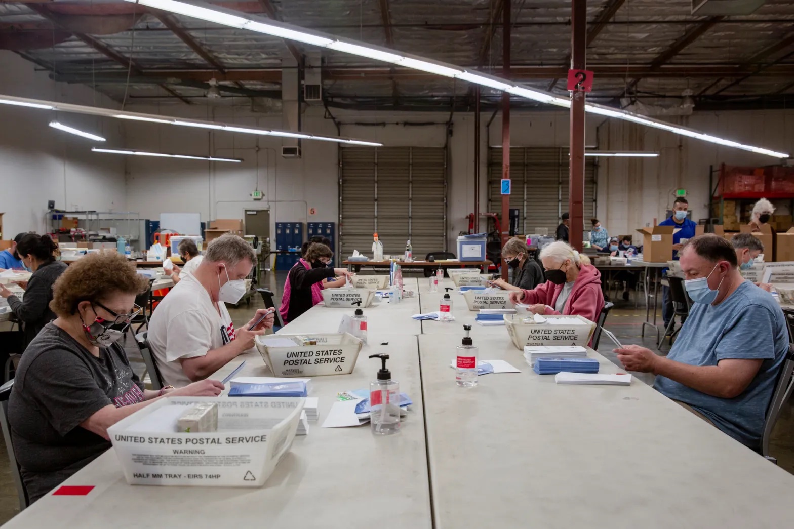 people sit space out at a long folding table in a warehouse, with stacks of envelopes and bins in front of them