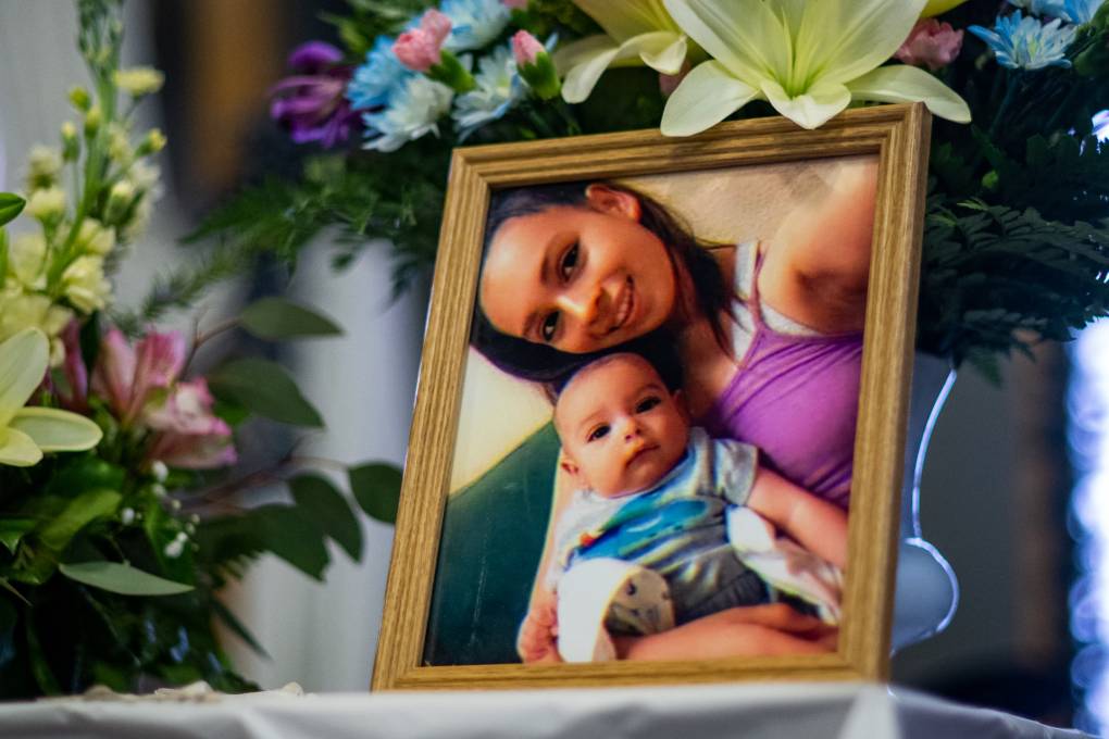 A framed photograph of a young mother smiling as she holds her baby sits on top of an altar honoring the two at their funeral service. Bouquets of blue, yellow and pink flowers surround the wooden frame.