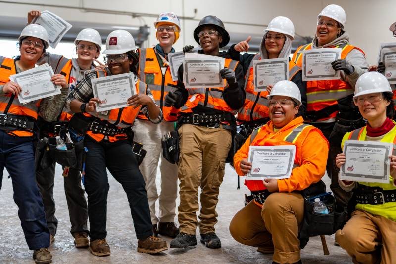 A group of people wearing construction uniforms pose holding certificates in a building.