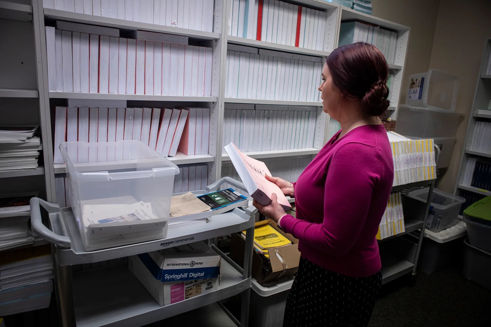 the side view of a middle-aged woman with her hair pulled back, filing a stack of white books on a shelf