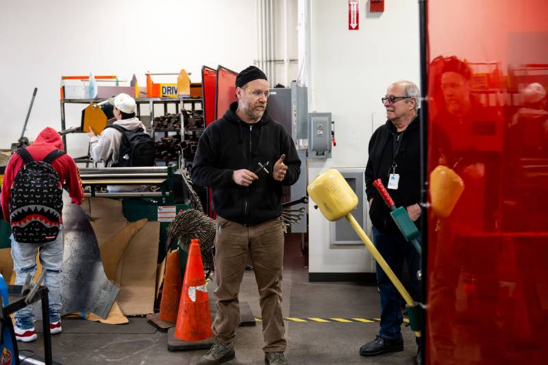 A white man with glasses in a black hoodie talks to another white man in a black beanie hat and black hoodie in a welding classroom.