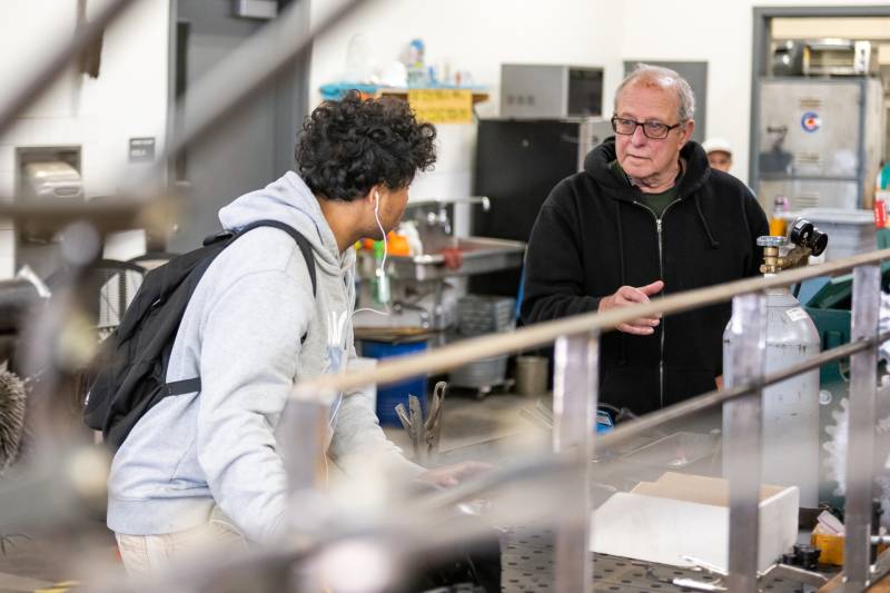 A white man with glasses in a black hoodie talks to a teenager wearing a gray hoodie with wired ear buds in his ear in a classroom.